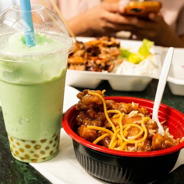 Bowl of orange chicken, noodles, and rice with a green boba smoothie on a restaurant table.