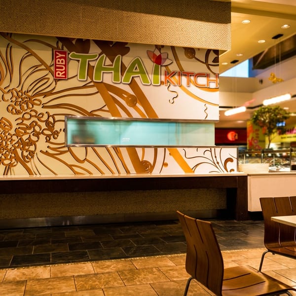 Ruby Thai Kitchen storefront with floral wall mural, illuminated counter, and wooden tables and chairs in a shopping-center dining area.