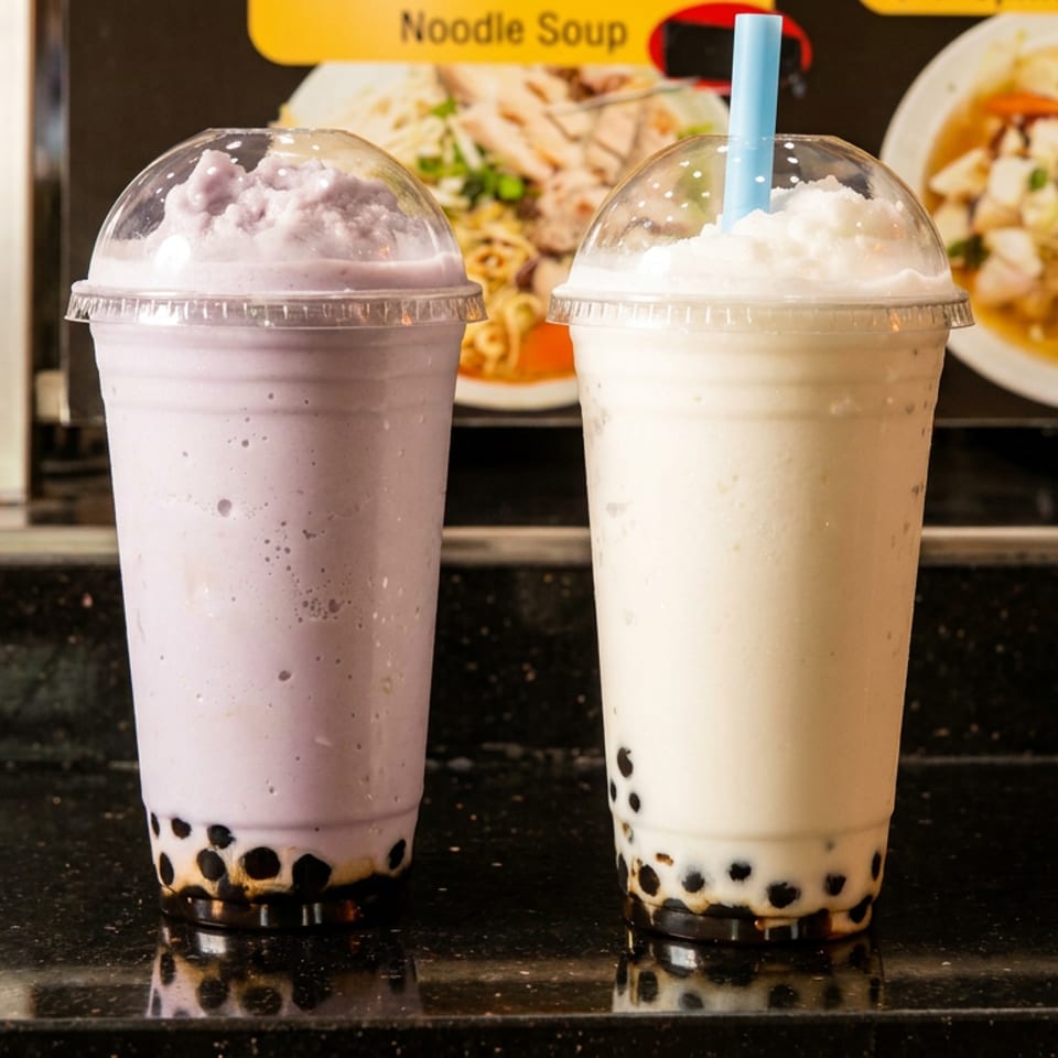 Two colorful bubble tea drinks with tapioca pearls sit on a restaurant counter, with noodle soup dishes pictured in the background.